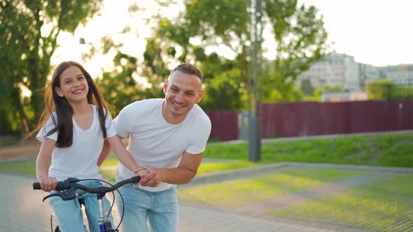Dad is Teaching Daughter How to Ride Bicycle at Sunset alt