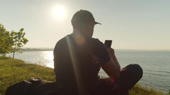 Man Tourist is Resting on the Seashore with a Smartphone in His Hand alt