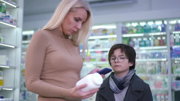 Curios Son Standing in Drugstore with Mother Checking Medication Online Using Smartphone alt