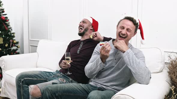 Playful gay couple blowing party horns while sitting on the couch celebrating Christmas