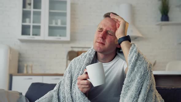 Sick young man with fever wrapped in warm blanket drinking hot tea alt