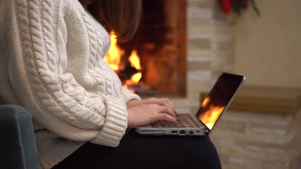 A Young Pregnant Woman is Sitting in an Armchair By the Fireplace and Typing on a Laptop alt