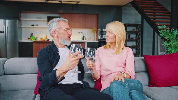 Happy Middle Aged Adult Married Couple Drinking White Wine From Glasses While Sitting on Sofa alt