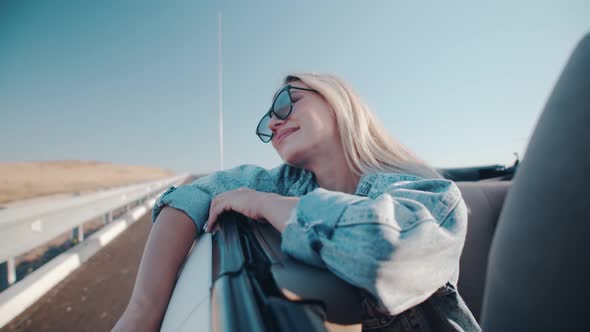 In the Back Seat of a Cabriolet, a Beautiful Woman Admires the landscape.A Young Woman in a Denim alt