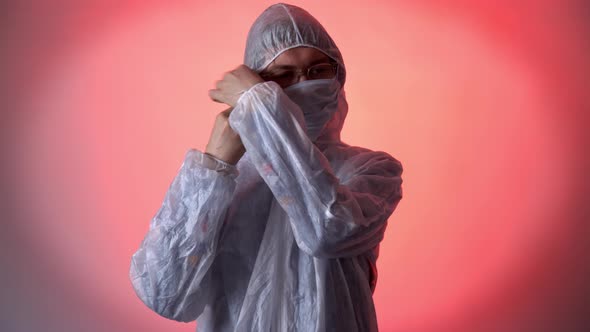 Serious Man in a White Protective Suit on Red Background Puts Medicine Mask, Zips Jacket, Puts on a