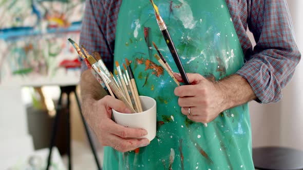 Closeup of Artist's Male Hands Holding Brushes at Level of Abdomenapron Stained with Paint alt