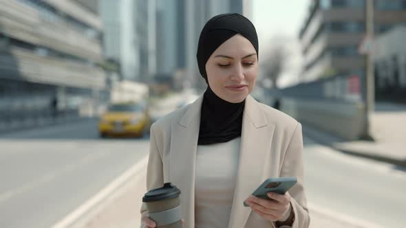 Woman in Hijab Using Mobile and Drinking Coffee Outdoors alt