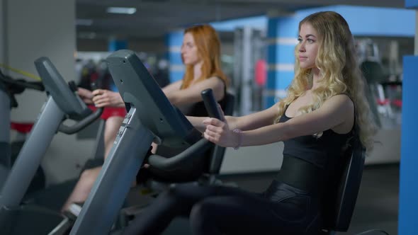 Side View of Confident Fit Blond Sportswoman and Redhead Woman at Background Riding Exercise Bikes alt
