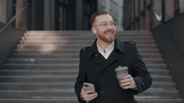 Businessman is Walking Near the Business Center with a Cup of Coffee and Smartphone in His Hand alt
