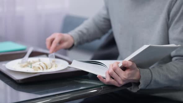Unrecognizable Caucasian Young Man Reading Book Eating Rolled Stuffed Pancakes in Canteen Indoors alt