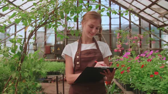 Female Gardener Checks Quality of Plants Takes Notes on Digital Tablet Walking Along Garden Center