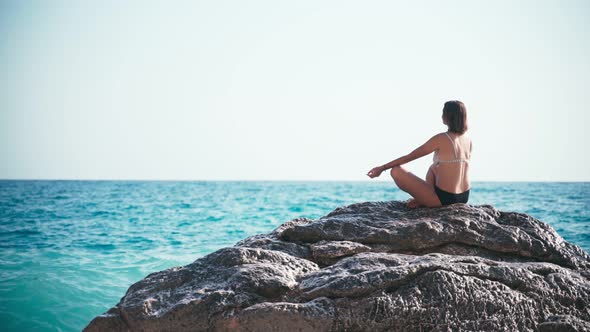 A Young Woman Sitting on a Big Rock at the Edge of the Sea and Meditating alt