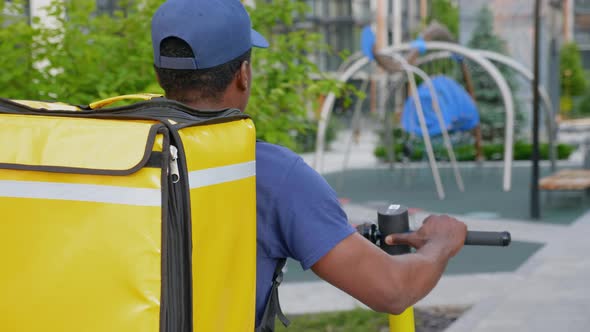 African Man Courier with Thermal Backpack Walks Street with Electric Scooter alt