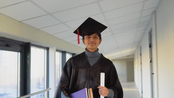 Pakistani Graduate in Mantle Stands with a Diploma and Books in Her Hands and Smiles alt