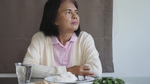 Sad senior Asian woman sitting in dining room with plenty of food, looking away. alt