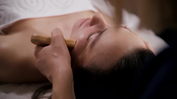 Massage Session  Young Woman Therapist Using Wooden Piece on the Cheek of Her Client alt