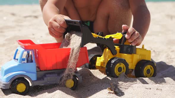 Close-up of a Boy's Tanned Hands Playing on the Beach with Cars, a Plastic Toy Car, an Excavator alt