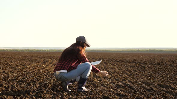 A Farmer Checks Quality of the Soil Before Sowing. Woman Farmer with a Tablet in Field Holds Earth alt