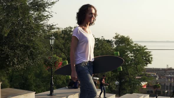 Young Attractive Girl Walking Holding Her Longboard in the City Street During Sunrise Slow Motion alt