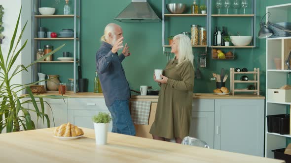 Elderly Couple Relaxing at Home Talking Over a Cup Of Tea in a Cozy Kitchen alt