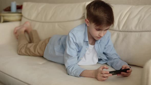 Child Lies on the Couch in His Hands Holding a Phone Plays with It