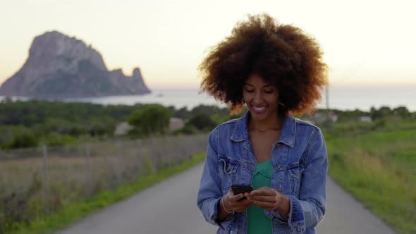 Smiling woman using smartphone on road at sunset alt