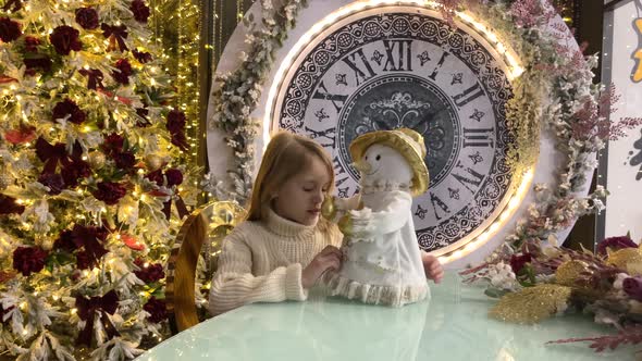 A Child Girl Play with a Doll Snowman in Near a Christmas Tree with Big Clock alt