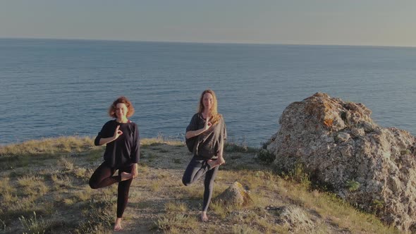 Women in Sportswear and Barefoot Do Yoga Exercises Against the Backdrop of the Majestic Coastal alt