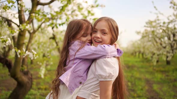 Two Girls Sisters Have Fun and Cuddle in the Garden with Flowering Trees alt