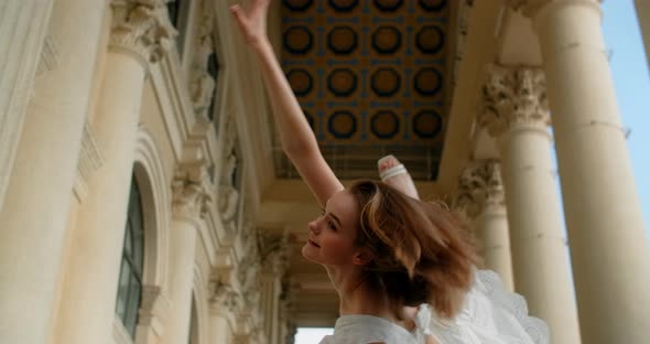 Young Balerina in White Dress are Doing Reverse Leg Swings Between the Columns of the Theater alt