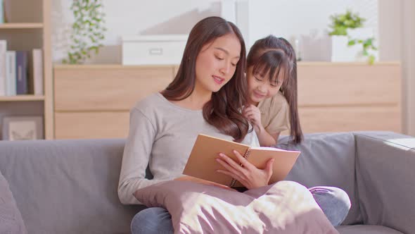An Asian mom and her kid are reading together. Mom is teaching her child alt