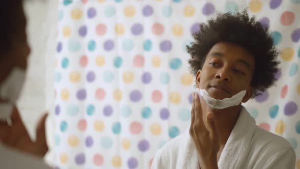 Black Guy in Bathrobe Applying Shave Cream on Face Standing Near Mirror in Bathroom alt