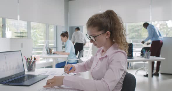 Side View of Young Businesswoman Working on Laptop and Paperwork in Office alt