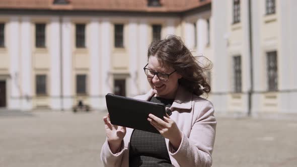 Happy Pretty Woman in Glasses Uses Mobile Guide in Tablet to Explore Landmark alt