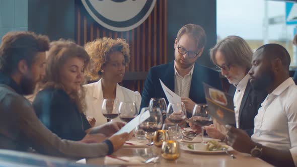 Group of Diverse Cheerful Friends Drinking Wine and Reading Menu in Modern Restaurant alt
