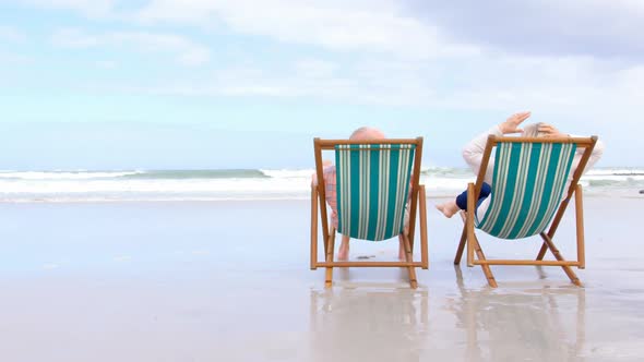 Rear view of old caucasian senior couple sitting on sun lounger and holding hands at beach 4kRear vi alt