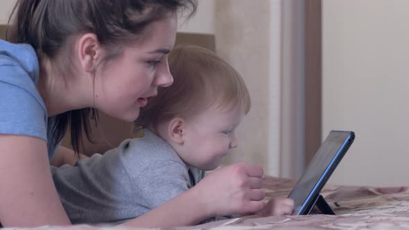 Modern Family Leisure, Nice Infant Boy with Young Mum Have Fun with Touch Tablet Lying on Bed at alt