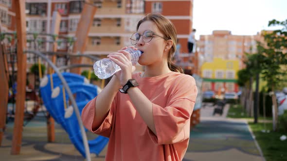 A Teenage Girl with Glasses Drinks Water From a Plastic Bottle on the Playground