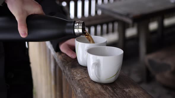 White male hand pouring tea from a thermos flask into two white cups in wooden cabin. alt