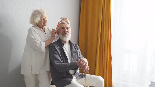 Smiling Senior Wife Caring for Husband Combing His Hair at Home