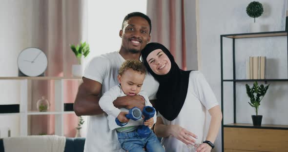 African Man and Muslim Woman and their Dark-Skinned Small Child which Looking at Camera at Home alt