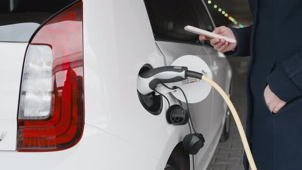 Woman Unplugging the Power Supply Cable From an Electric Car and Using Her Smartphone for Unlocking alt