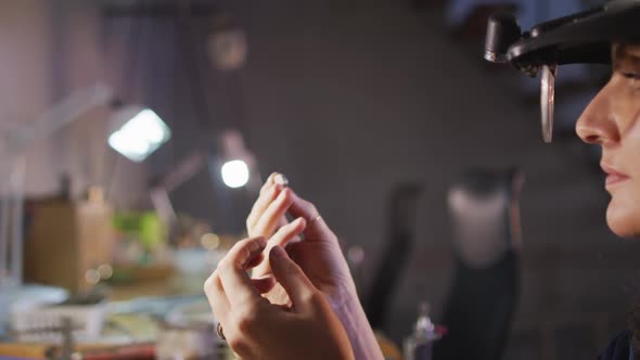 Close up of hands of caucasian female jeweller wearing glasses, checking jewelry at workshop alt