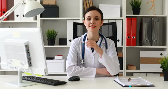 Doctor Sitting at Workplace Raising Hand To Chin and Looking at Camera