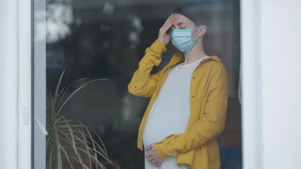 Stressed Worried Pregnant Woman in Covid19 Face Mask with Head Ache Standing Indoors Looking Out alt
