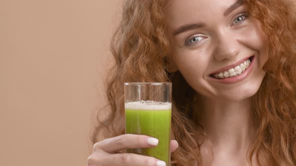 Happy RedHaired Lady Holding Green Smoothie Glass Over Beige Background alt