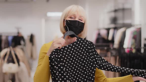 Happy Mature Woman in Protective Mask in Clothing Store Joyful Female Customer Shopaholic Trying on alt