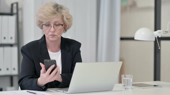 Old Businesswoman Using Smartphone While Working on Laptop alt