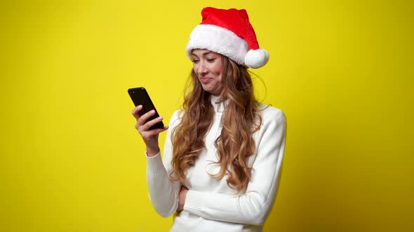 Woman Christmas Santa Hat White Sweater Yellow Studio Background with Smartphone in Hand alt