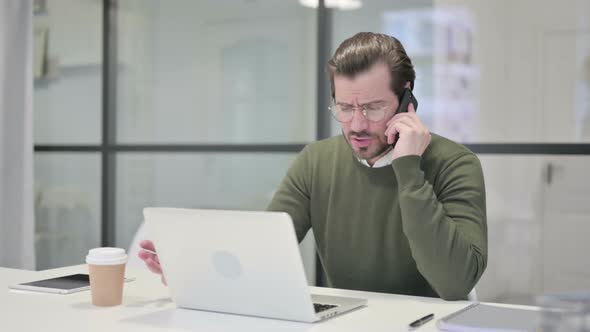 Angry Young Businessman Talking on Smartphone While Using Laptop in Office alt